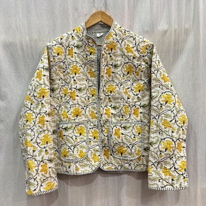 Pode incluir: Casaco acolchoado com padrão floral em amarelo e verde sobre fundo branco. O casaco tem gola alta, zíper inteiro e dois bolsos frontais. As bordas são adornadas com um tecido listrado em preto e branco.