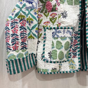 Pu&ograve; includere: Una giacca trapuntata bianca con un bordo a righe verde e bianco. La giacca ha un design patchwork con motivi floreali e geometrici in tonalit&agrave; di rosa, verde e blu.