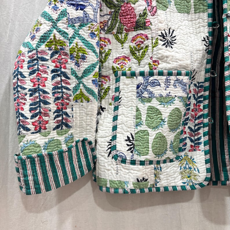 Pu&ograve; includere: Una giacca trapuntata bianca con un bordo a righe verde e bianco. La giacca ha un design patchwork con motivi floreali e geometrici in tonalit&agrave; di rosa, verde e blu.