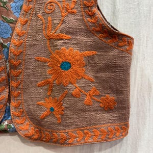 K&ouml;nnte beinhalten: Nahaufnahme einer braunen Weste mit orangefarbener Blumenstickerei. Die Weste hat einen strukturierten Stoff und ein Blumendesign mit einem t&uuml;rkisfarbenen Zentrum. Die Kanten sind mit orangefarbenen, blattartigen Mustern verziert. Die Weste ist Teil eines gr&ouml;&szlig;eren Kleidungsst&uuml;cks.