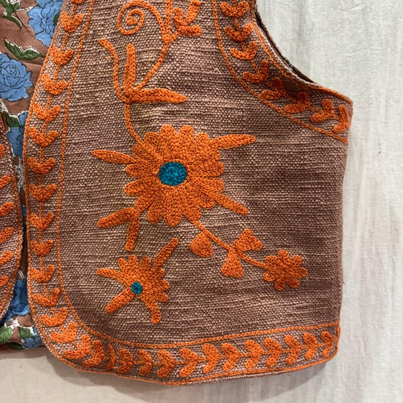 K&ouml;nnte beinhalten: Nahaufnahme einer braunen Weste mit orangefarbener Blumenstickerei. Die Weste hat einen strukturierten Stoff und ein Blumendesign mit einem t&uuml;rkisfarbenen Zentrum. Die Kanten sind mit orangefarbenen, blattartigen Mustern verziert. Die Weste ist Teil eines gr&ouml;&szlig;eren Kleidungsst&uuml;cks.