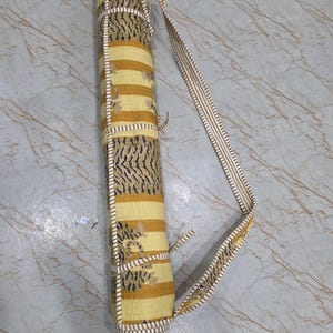 May include: A cylindrical yoga mat bag featuring a yellow and white striped pattern with black tiger-inspired motifs. The bag includes a long, adjustable shoulder strap with matching white and yellow stripes and thin fabric ties for secure closure.