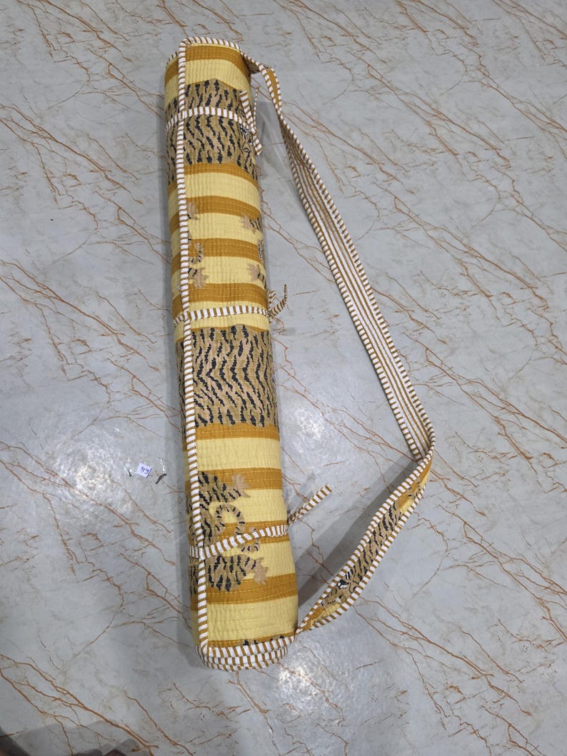 May include: A cylindrical yoga mat bag featuring a yellow and white striped pattern with black tiger-inspired motifs. The bag includes a long, adjustable shoulder strap with matching white and yellow stripes and thin fabric ties for secure closure.