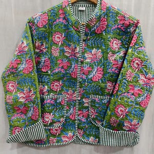 Puede incluir: Chaqueta acolchada verde con un estampado floral en rosa y azul. La chaqueta tiene cuello mandarín, dos bolsillos y un ribete a rayas. La chaqueta está colgada en una percha de madera.