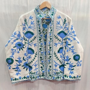 Puede incluir: Chaqueta color crema con intrincado bordado floral azul y verde. La chaqueta presenta un cuello mandarín y se muestra en una percha de madera. El bordado crea un efecto decorativo texturizado, realzando el atractivo visual de la chaqueta.
