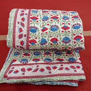 Könnte beinhalten: Eine gefaltete, gesteppte Decke mit einem Blumenmuster in Rot und Blau auf weißem Hintergrund. Die Decke hat einen Rand mit einem sich wiederholenden Blumendesign und ist auf einer roten Oberfläche gefaltet.