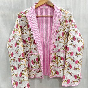 May include: A quilted jacket with a floral pattern in pink and green on a white background. The jacket has a pink striped lining and trim, with two front pockets and wooden buttons. The jacket is hanging on a wooden hanger.