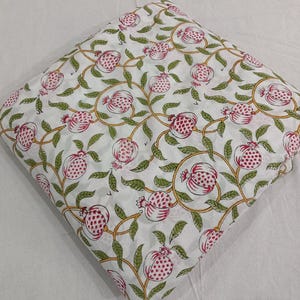 May include: A folded piece of white fabric featuring a repeating pattern of pink pomegranate fruits with green leaves and yellow stems. The design is arranged in a continuous botanical motif across the entire surface of the textile material.