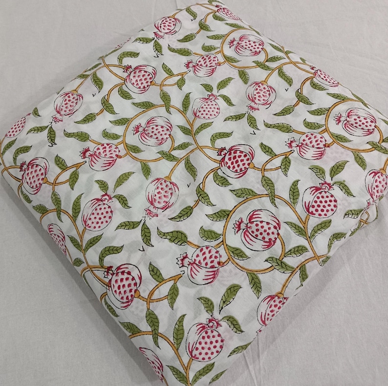 May include: A folded piece of white fabric featuring a repeating pattern of pink pomegranate fruits with green leaves and yellow stems. The design is arranged in a continuous botanical motif across the entire surface of the textile material.