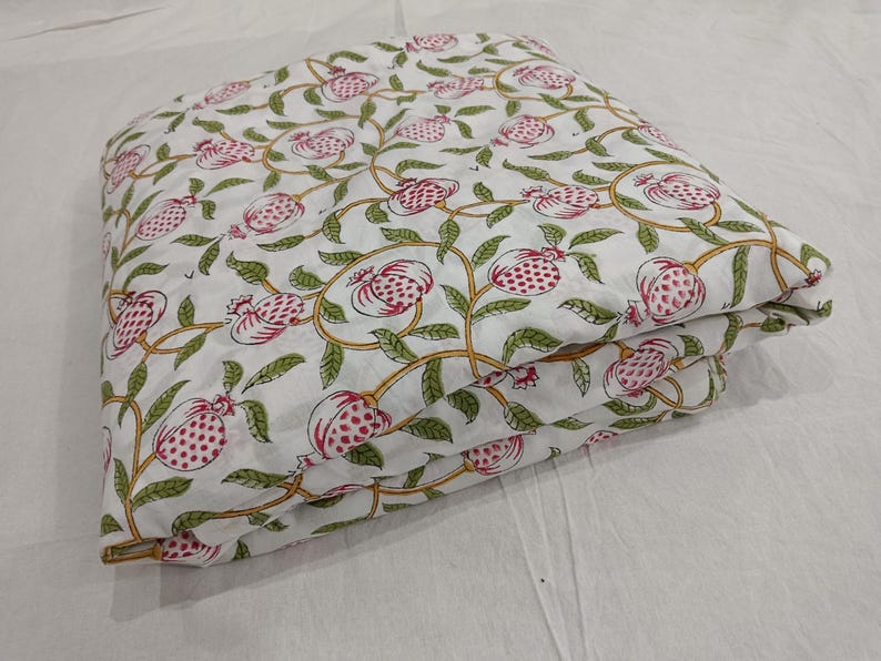 May include: A folded piece of white fabric featuring a repeating pattern of pink pomegranates with green leaves and yellow stems. The textile displays a botanical print with stylized fruit and vine motifs arranged in a continuous, flowing design.