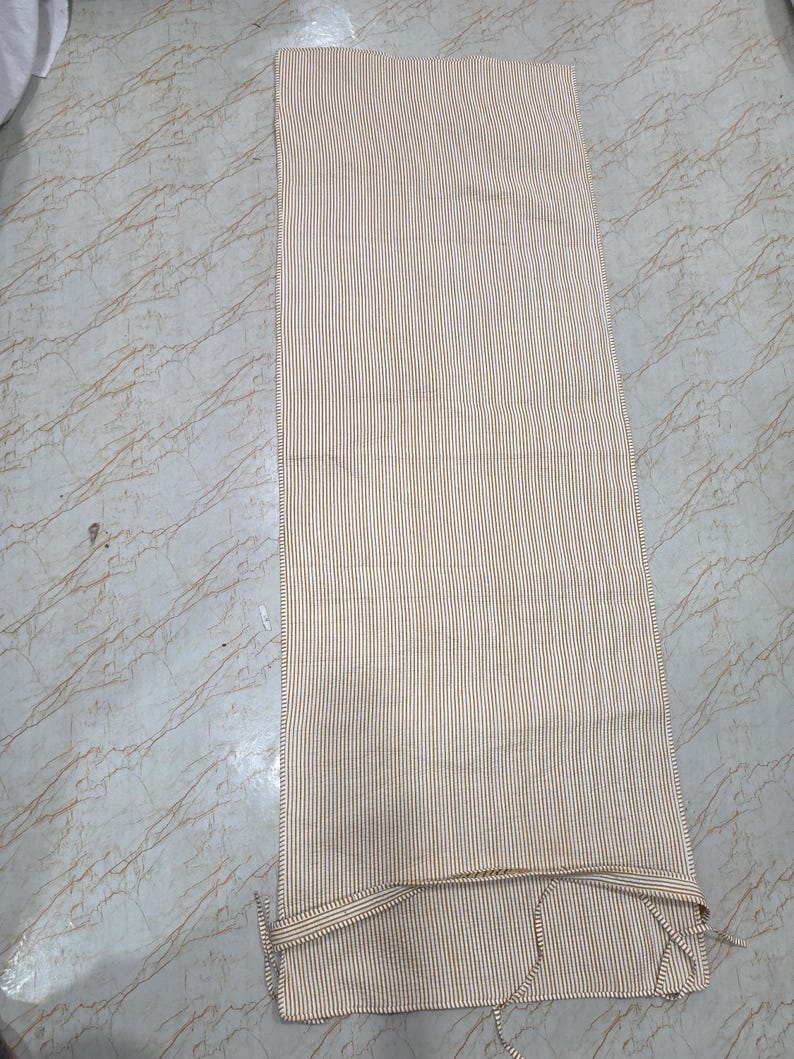 May include: A rectangular fabric runner featuring a fine vertical pinstripe pattern in mustard yellow and white. The textile has finished edges and includes thin fabric ties at the bottom corners for securing the piece in place during use.