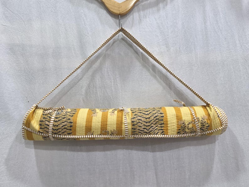 May include: A rolled fabric mat featuring a repeating pattern of yellow and white vertical stripes interspersed with sections of dark tiger-print motifs. The mat is secured with a matching striped fabric strap and hangs from a wooden hanger.