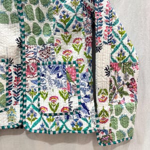Pu&ograve; includere: Una giacca trapuntata bianca con un design a patchwork che presenta motivi floreali e geometrici in tonalit&agrave; di blu, verde, rosa e bianco. La giacca ha una chiusura con bottone e una tasca sul lato sinistro.