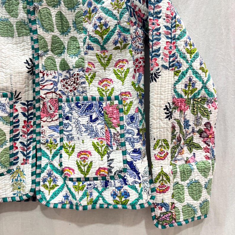 Pu&ograve; includere: Una giacca trapuntata bianca con un design a patchwork che presenta motivi floreali e geometrici in tonalit&agrave; di blu, verde, rosa e bianco. La giacca ha una chiusura con bottone e una tasca sul lato sinistro.