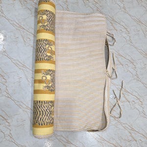 May include: A quilted fabric roll featuring a yellow and white striped pattern with a repeating dark gray tiger print on one side and a fine white and yellow pinstripe pattern on the other, complete with thin fabric ties for securing the roll.