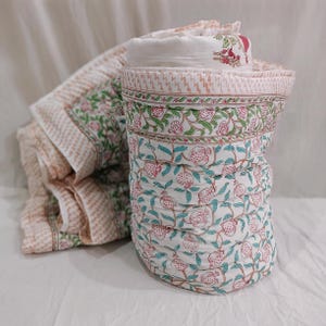 Könnte beinhalten: Eine gerollte und eine ausgebreitete Steppdecke mit floralem Muster. Die Decke zeigt ein sich wiederholendes Muster aus rosa Granatäpfeln, grünen Blättern und blauen Ranken auf weißem Grund. Der Rand hat ein rosa-weißes Zickzackmuster.