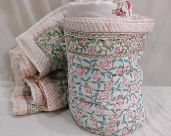May include: A rolled-up, white and floral-patterned quilt with a matching unfolded section. The quilt features a repeating pattern of pink pomegranates, green leaves, and blue vines on a white background. The border has a pink and white zigzag design.
