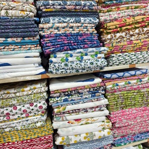 May include: Multiple stacks of folded cotton fabric featuring a variety of colorful block-printed patterns. The textiles display diverse floral, geometric, and botanical motifs in shades of blue, pink, yellow, green, and red on white backgrounds.
