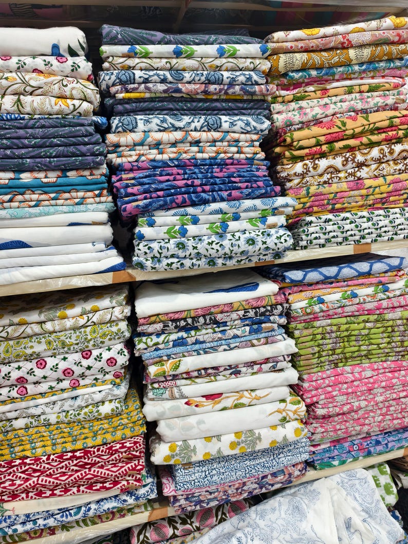 May include: Multiple stacks of folded cotton fabric featuring a variety of colorful block-printed patterns. The textiles display diverse floral, geometric, and botanical motifs in shades of blue, pink, yellow, green, and red on white backgrounds.