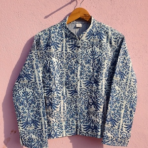 May include: A blue and white quilted jacket with a botanical pattern, hanging on a wooden hanger against a pink wall. The jacket features a mandarin collar and a button-up front. The pattern includes leaf and floral motifs.