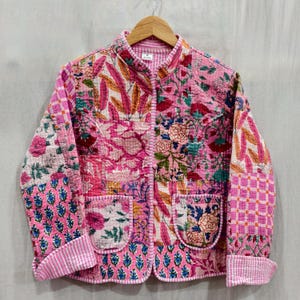 Puede incluir: Chaqueta acolchada rosa con estampado floral. La chaqueta tiene cuello mandarín, dos bolsillos delanteros y cierre de botones. El diseño floral incluye flores y hojas rosas, rojas y azules. La chaqueta está colgada en una percha de madera.