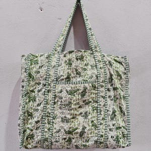 Puede incluir: Un bolso tote blanco con un diseño de estampado de animales verdes. El bolso tiene forma rectangular con un bolsillo frontal y dos asas. Los bordes y las asas están ribeteados con un borde verde claro.