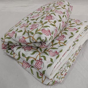 May include: A folded white fabric featuring a repeating pattern of pink pomegranates with green leaves and yellow stems. The textile is layered in multiple stacks, showcasing the botanical print across the surface of the soft, light material.