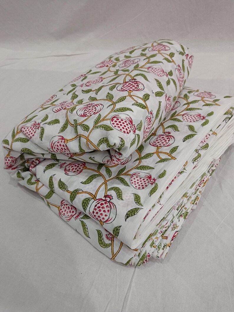May include: A folded white fabric featuring a repeating pattern of pink pomegranates with green leaves and yellow stems. The textile is layered in multiple stacks, showcasing the botanical print across the surface of the soft, light material.
