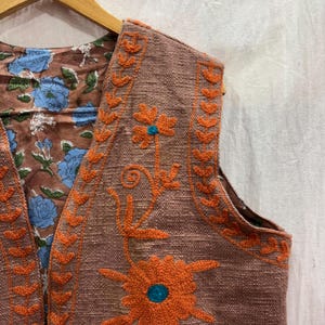 K&ouml;nnte beinhalten: Eine braune Weste mit orangefarbener Blumenstickerei und einem blauen Zentrum. Das Innere der Weste zeigt ein Blumenmuster mit blauen Blumen auf braunem Hintergrund. Die West ist an einem Holzb&uuml;gel aufgeh&auml;ngt.