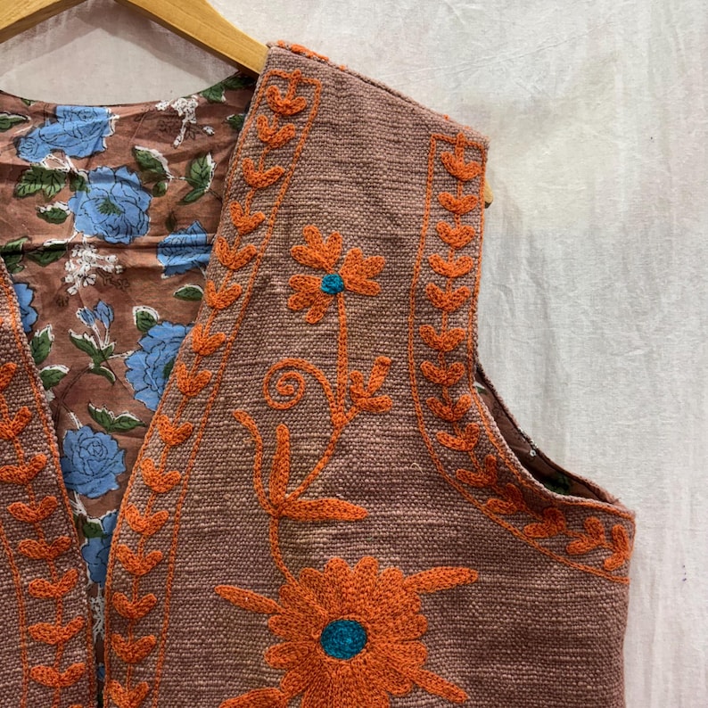 K&ouml;nnte beinhalten: Eine braune Weste mit orangefarbener Blumenstickerei und einem blauen Zentrum. Das Innere der Weste zeigt ein Blumenmuster mit blauen Blumen auf braunem Hintergrund. Die West ist an einem Holzb&uuml;gel aufgeh&auml;ngt.