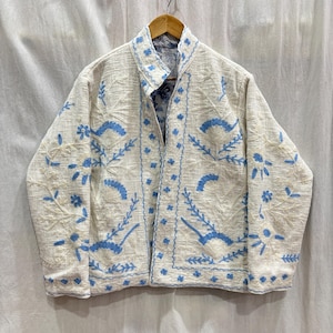 Puede incluir: Chaqueta color crema con bordado floral azul. La chaqueta abierta presenta una tela texturizada y un estampado floral azul contrastante en el forro interior. El bordado incluye flores y patrones de hojas estilizados.