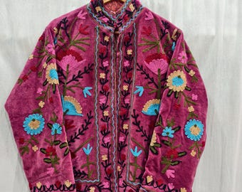 May include: A magenta velvet jacket with intricate floral embroidery in shades of pink, blue, and green. The jacket features a high collar and a button-up front. The embroidery includes flowers, leaves, and other botanical elements.