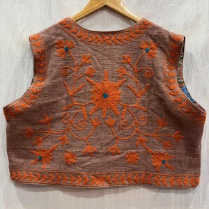 K&ouml;nnte beinhalten: Eine &auml;rmellose Weste in Braun mit aufwendiger orangefarbener Blumenstickerei. Das Design zeigt eine gro&szlig;e zentrale Blume und kleinere Bl&uuml;ten mit t&uuml;rkisfarbenen Akzenten. Die Kanten sind ebenfalls orange bestickt, was einen stimmigen Look erzeugt.