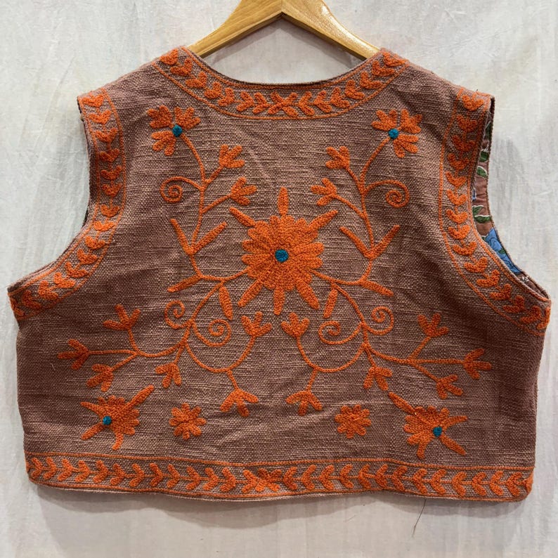 K&ouml;nnte beinhalten: Eine &auml;rmellose Weste in Braun mit aufwendiger orangefarbener Blumenstickerei. Das Design zeigt eine gro&szlig;e zentrale Blume und kleinere Bl&uuml;ten mit t&uuml;rkisfarbenen Akzenten. Die Kanten sind ebenfalls orange bestickt, was einen stimmigen Look erzeugt.