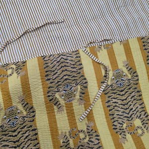 May include: A quilted textile featuring a yellow and white vertical striped pattern on the top section and a yellow background with repeating black and white tiger illustrations on the bottom section, complete with thin matching striped fabric ties.
