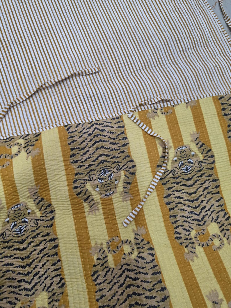 May include: A quilted textile featuring a yellow and white vertical striped pattern on the top section and a yellow background with repeating black and white tiger illustrations on the bottom section, complete with thin matching striped fabric ties.