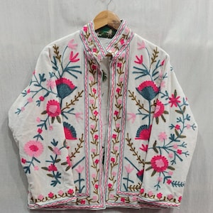 May include: Cream-coloured jacket with detailed floral embroidery in pink, teal, and green. The jacket has a stand-up collar and is decorated with intricate stitching along the edges and seams. The floral design includes various flower and leaf patterns.