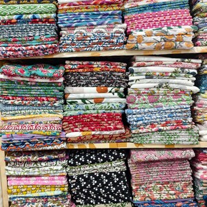 May include: Multiple stacks of folded cotton fabric arranged on wooden shelves. The textiles feature a wide variety of colorful patterns, including floral, geometric, and abstract prints in shades of pink, blue, green, red, yellow, and black.