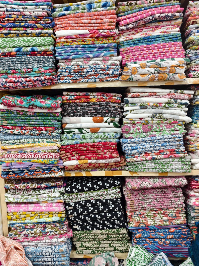 May include: Multiple stacks of folded cotton fabric arranged on wooden shelves. The textiles feature a wide variety of colorful patterns, including floral, geometric, and abstract prints in shades of pink, blue, green, red, yellow, and black.