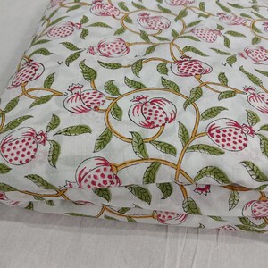 May include: A folded piece of white fabric featuring a repeating pattern of pink pomegranates with red seeds, green leaves, and yellow vines. The textile displays a consistent botanical print across the surface, showcasing a detailed illustration of fruit and foliage arranged in a flowing, interconnected design.