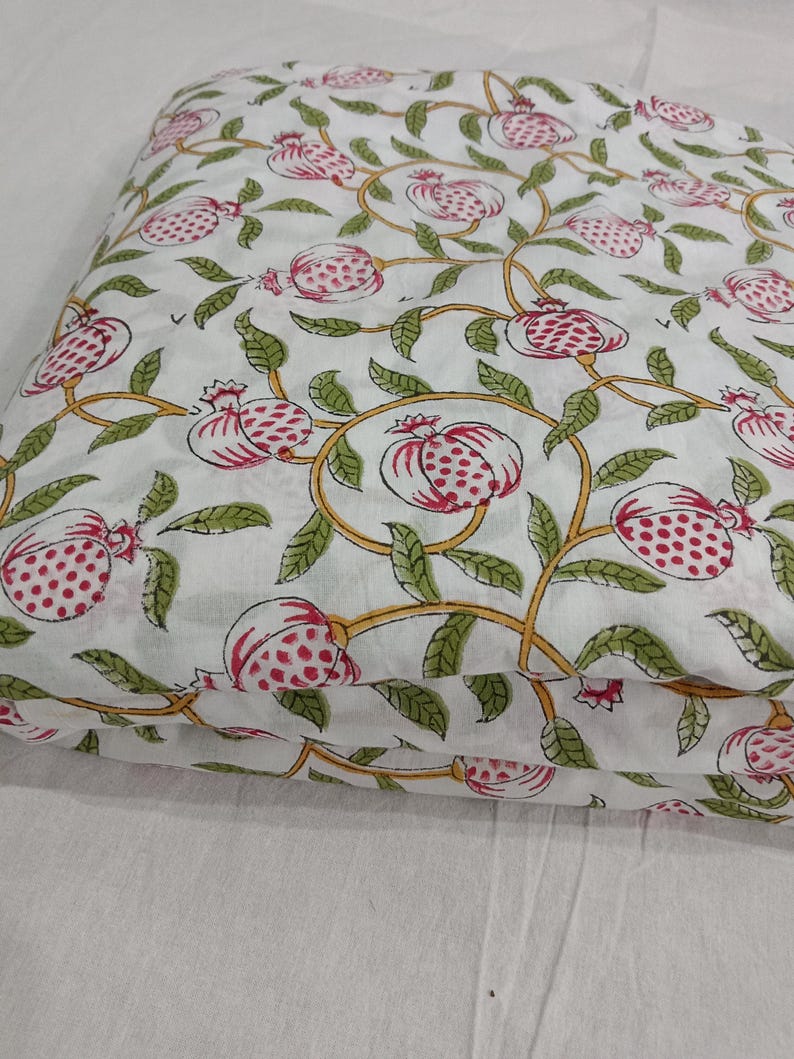 May include: A folded piece of white fabric featuring a repeating pattern of pink pomegranates with red seeds, green leaves, and yellow vines. The textile displays a consistent botanical print across the surface, showcasing a detailed illustration of fruit and foliage arranged in a flowing, interconnected design.