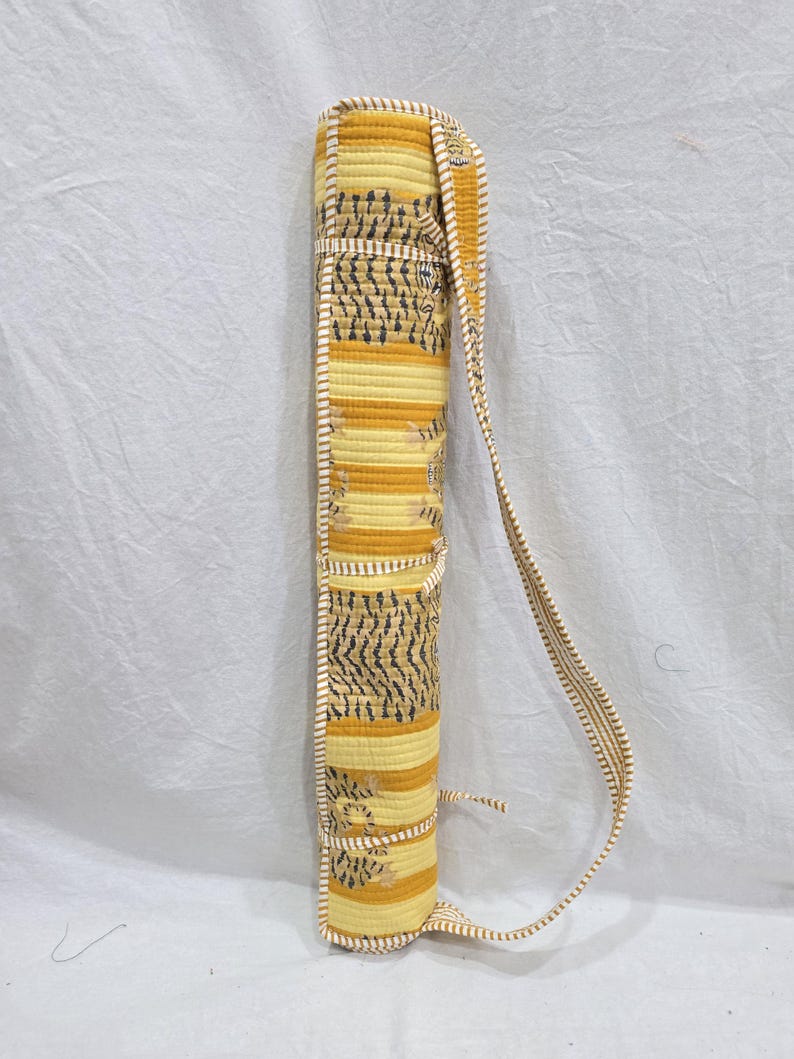 May include: A cylindrical quilted carrying case featuring a yellow and white horizontal striped pattern with dark gray tiger-like markings. The case includes a long, matching striped shoulder strap and a tie closure at the top and bottom edges.