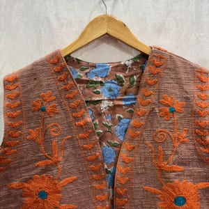 K&ouml;nnte beinhalten: Eine braune Weste mit orangefarbener Blumenstickerei und t&uuml;rkisfarbenen Akzenten. Die Weste hat ein floral gemustertes Futter mit blauen Blumen. Die Weste h&auml;ngt an einem Holzkleiderb&uuml;gel.