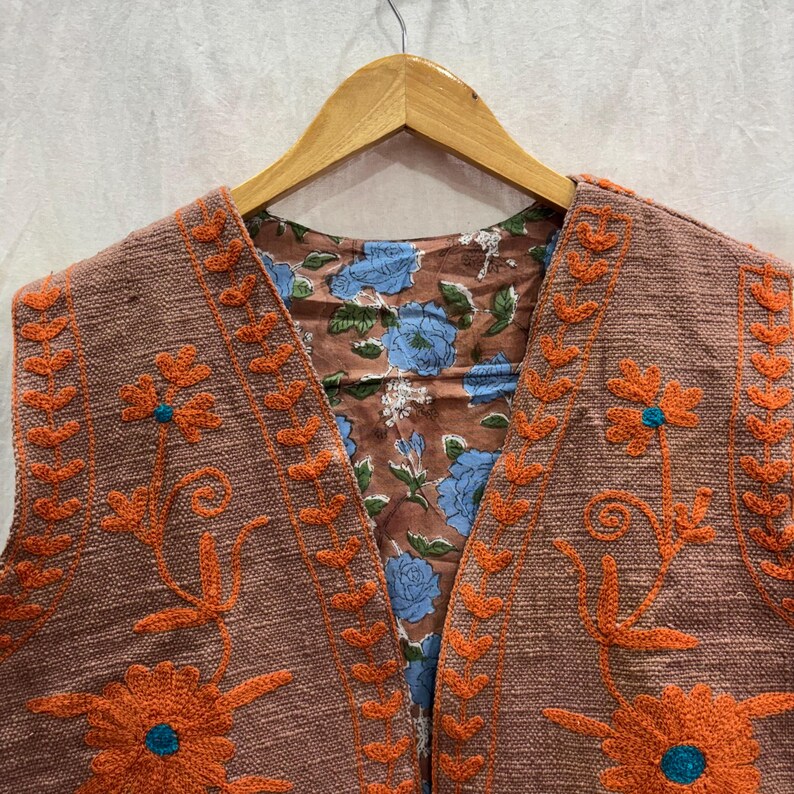 K&ouml;nnte beinhalten: Eine braune Weste mit orangefarbener Blumenstickerei und t&uuml;rkisfarbenen Akzenten. Die Weste hat ein floral gemustertes Futter mit blauen Blumen. Die Weste h&auml;ngt an einem Holzkleiderb&uuml;gel.