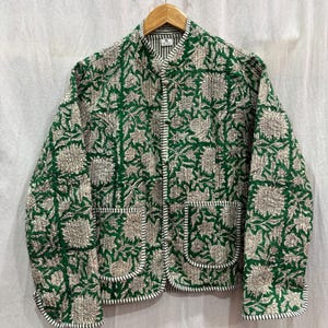 Pode incluir: Casaco acolchoado verde com um padrão floral em creme e branco. O casaco tem gola mandarim, dois bolsos frontais e é adornado com tecido listrado branco e preto. Está pendurado num cabide de madeira.