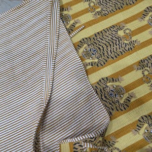 May include: A reversible quilted textile featuring a pattern of tigers on a yellow and mustard striped background on one side, and a fine white and mustard striped pattern on the reverse side. The fabric has a visible stitched texture throughout.