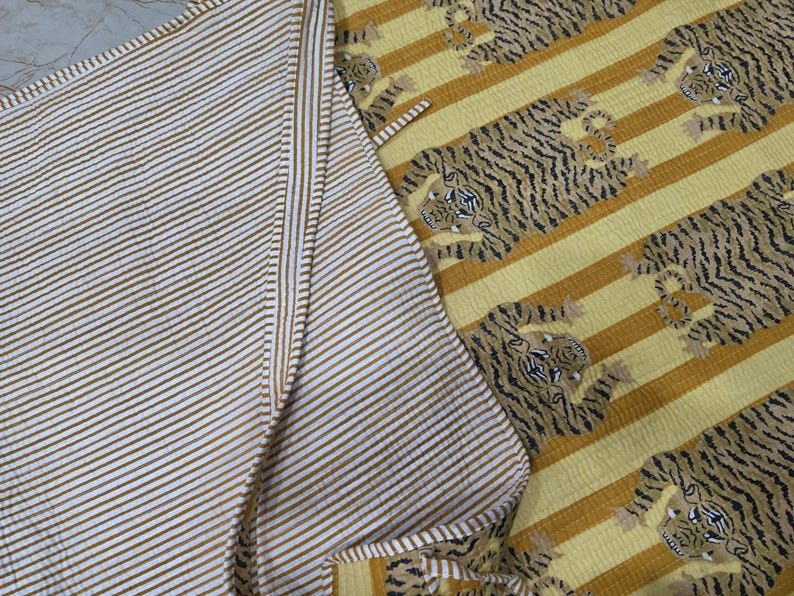 May include: A reversible quilted textile featuring a pattern of tigers on a yellow and mustard striped background on one side, and a fine white and mustard striped pattern on the reverse side. The fabric has a visible stitched texture throughout.