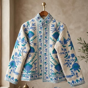 Könnte beinhalten: Eine cremefarbene Jacke mit Stehkragen und langen Ärmeln, verziert mit aufwendigen Stickereien in Blau- und Türkistönen. Die Blumen- und Rankenmuster sind symmetrisch auf den Vorderteilen und den Ärmeln angeordnet.