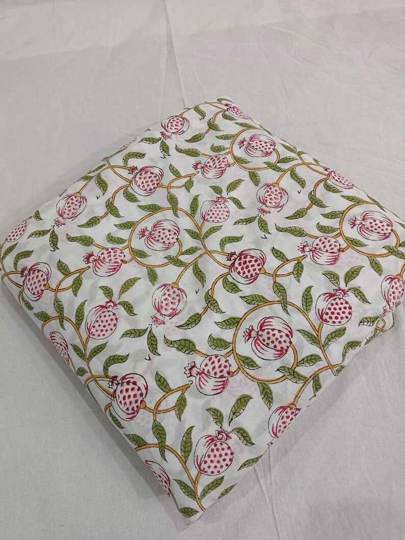 May include: A folded piece of white fabric featuring a repeating pattern of pink pomegranates with green leaves and yellow vines. The textile displays a block-printed style design with consistent spacing between the botanical motifs across the surface.