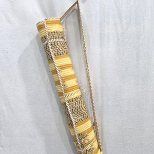 May include: A cylindrical yoga mat bag featuring a quilted texture with alternating horizontal bands of yellow, gold, and white stripes, accented by dark gray leaf-like patterns. The bag includes a long, thin, matching fabric shoulder strap.